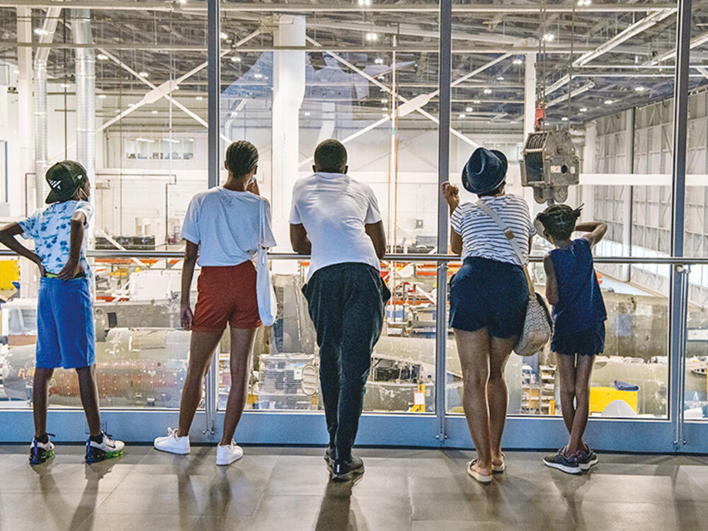 A group of five young visitors stand before the vast glass window at the second-floor overlook that offers an unfettered view of the Mary Baker Engen Restoration Hangar where aircraft sits ready for museum professionals to work on it in a workshop like atmosphere.