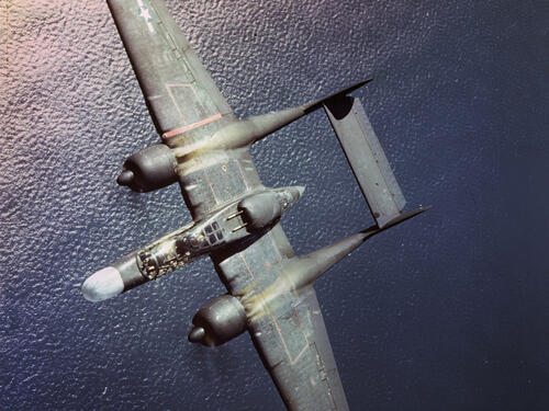 A gray, twin-engine military airplane with a twin-boom tail flies low over a dark ocean.